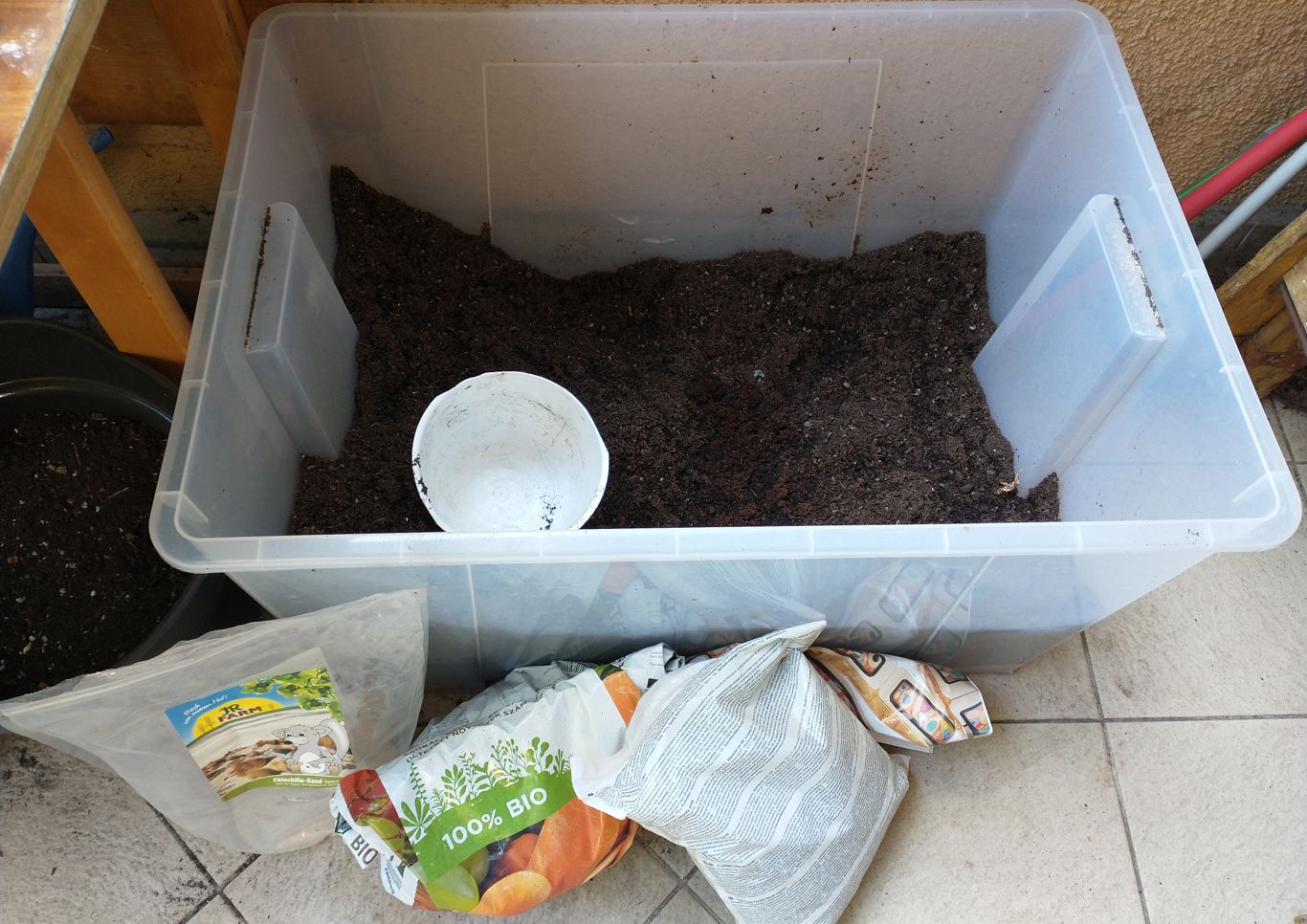 A plastic storage container with soil in it; beside it are a bag of sand, compost, and perlite.